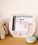 A portable white noise machine with a white base rests on a wooden surface next to a printed What to Wear guide showing room temperatures and appropriate sleepwear.