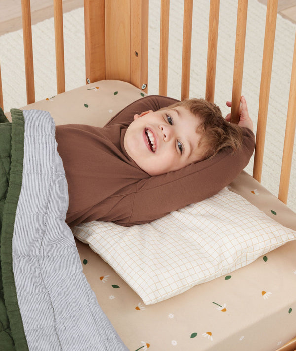 Toddler lying in a crib smiling using ergoPouch Organic Toddler Pillow Case in Caramel Grid