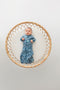 A baby lies on a round, woven rattan mat, wearing a blue zip-up swaddle sack.