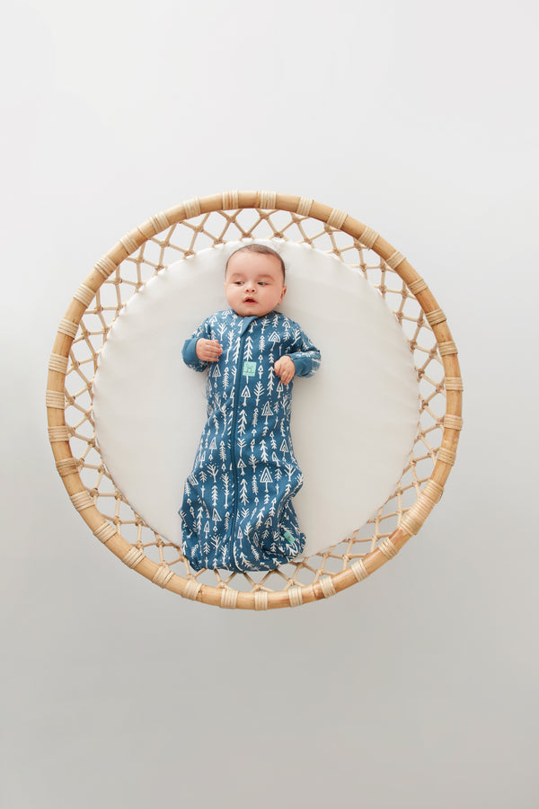 A baby lies on a round, woven rattan mat, wearing a blue zip-up swaddle sack.