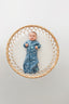 A baby lies on a round, woven rattan mat, wearing a blue zip-up swaddle sack.