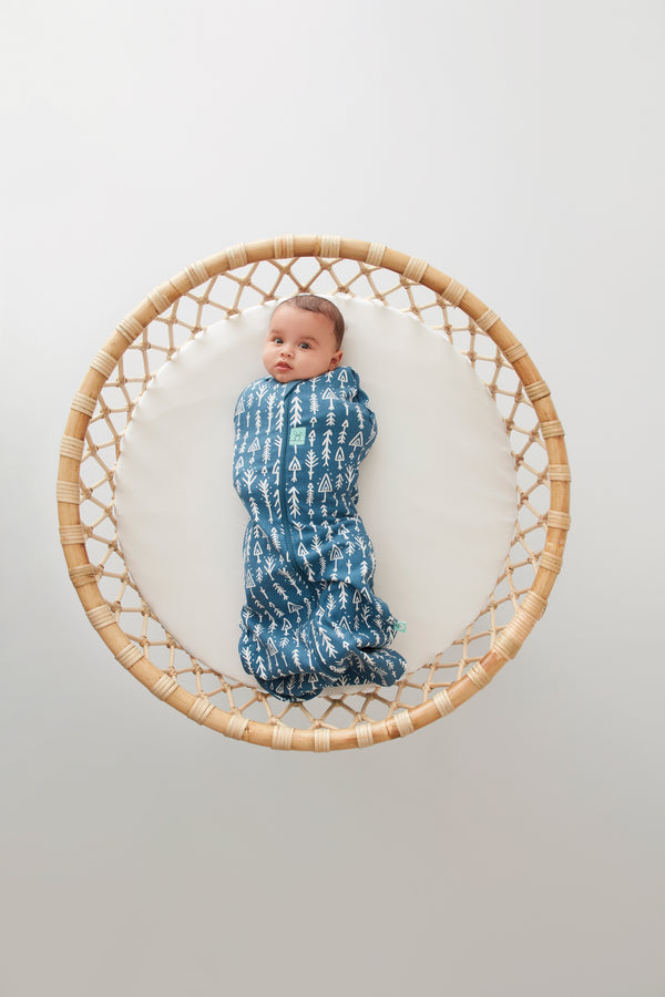 A baby lies on a round, woven rattan bassinet, wearing a blue zip-up cocoon swaddle sack