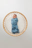 A baby lies on a round, woven rattan bassinet, wearing a blue zip-up cocoon swaddle sack
