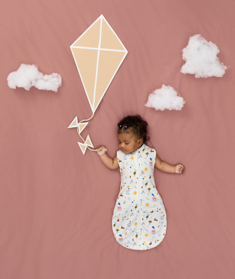 Newborn asleep in ergoPouch Cocoon Swaddle Sack 0.2 TOG Daydream, arms out sleeping, with a geometric kite and clouds