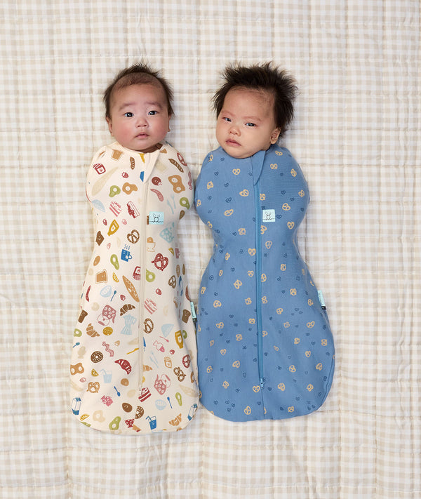 Two newborn babies lying side by side wearing ergoPouch Cocoon Swaddle Sack 0.2 TOG in Petit Pretzel and Bon Appetit