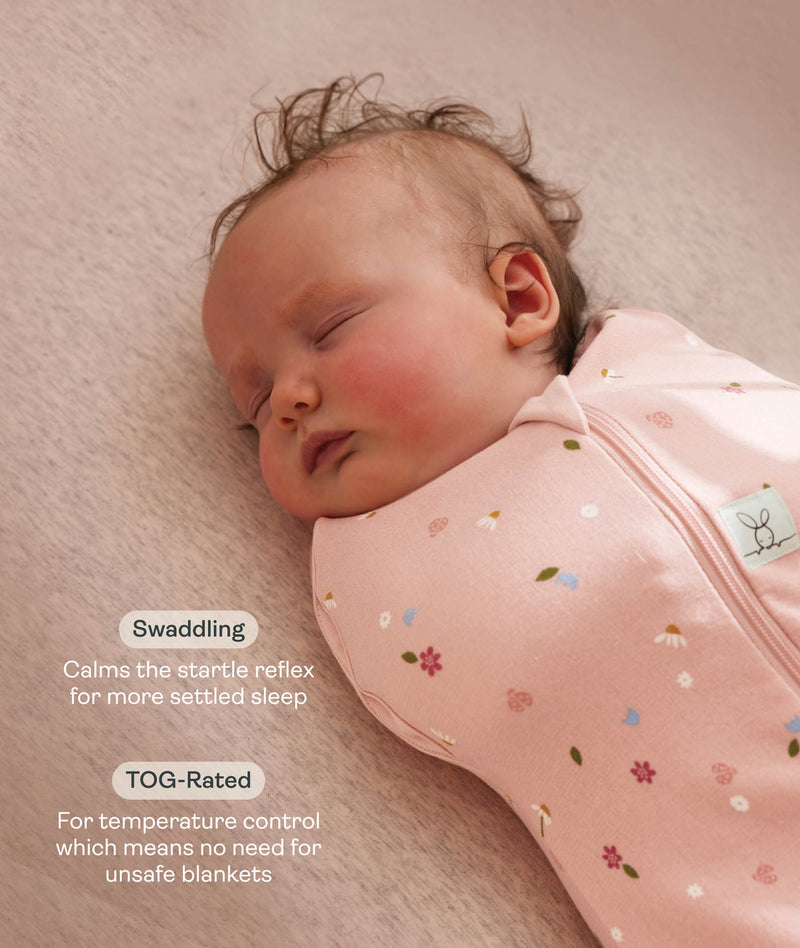 Close up of a newborn being swaddled in ergoPouch Cocoon Swaddle Sack 1.0 TOG Daisies with text highlighting swaddling benefit and TOG rating feature.