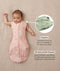 Baby lying on a beige surface in a pink floral swaddle sack with arms out, featuring callout text highlighting armhole poppers and a bell-shaped design.