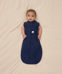Baby lying on a beige blanket wearing an ergoPouch Cocoon Swaddle Sack 1.0 TOG, transitioning to arms out sleeping.