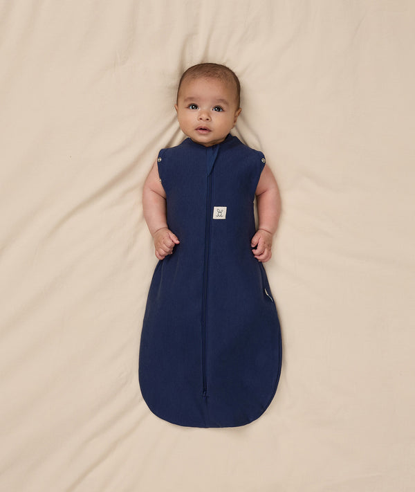 Baby lying on a beige blanket wearing an ergoPouch Cocoon Swaddle Sack 1.0 TOG, transitioning to arms out sleeping.