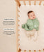 Baby lying in a wooden crib wearing a sage green patterned swaddle sack over footed pajamas, with product feature text callouts highlighting organic cotton fabric and two-way zipper features.
