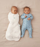 Two babies lying on their backs, one wearing an ergoPouch Cocoon Swaddle Sack 2.5 TOG Oatmeal Marle with arm in sleeping, and the other in a Long Sleeve Romper Dragonflies
