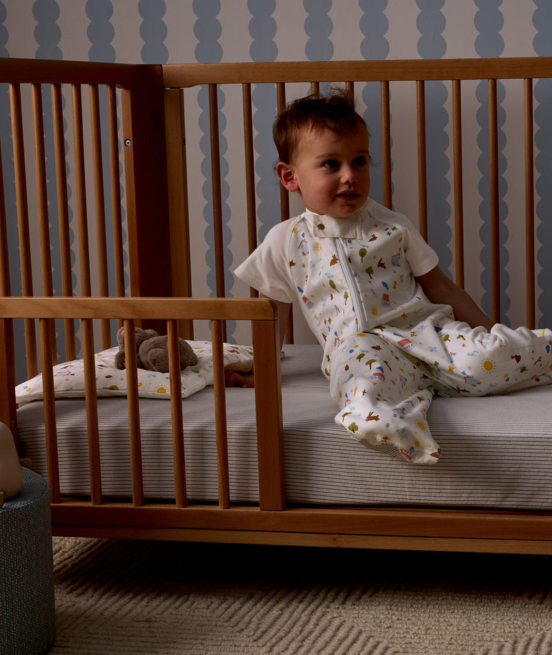 Child sitting in a wooden crib with Organic Toddler Pillow and Case Daydream, wearing a Sleep Suit Sack 1.0 TOG Daydream.