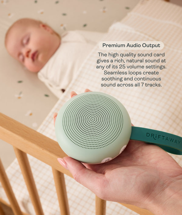 An adult hand holds a round, mint-green Drift Away white noise machine next to a crib where a baby is peacefully sleeping. A text box highlights 