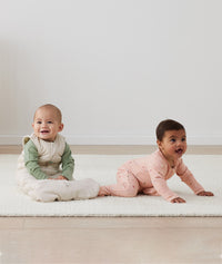 Two infants on the floor, one wearing an ergoPouch Long Sleeve Romper 1.0 TOG Daisies, and the other layered in a warmer Long Sleeve Romper 1.0 TOG Willow with a Sleep Sack 2.5 TOG Oatmeal Marle overtop for warmth. 