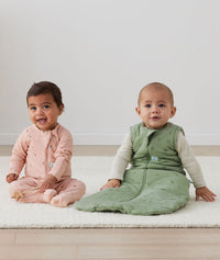 Two babies sitting on the floor, one wearing an ergoPouch Long Sleeve Romper 1.0 TOG Daisies, and the other layered in a warmer Long Sleeve Romper 1.0 TOG Oatmeal Marle with a Sleep Sack 2.5 TOG Willow overtop for warmth. 