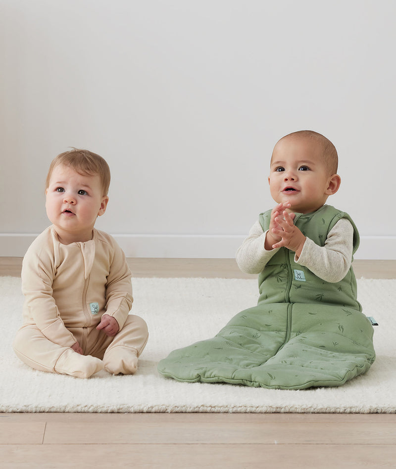 Two infants sitting on the floor, one wearing an ergoPouch Long Sleeve Romper 1.0 TOG Vanilla, and the other layered in a warmer Long Sleeve Romper 1.0 TOG Oatmeal Marle with a Sleep Sack 2.5 TOG Willow overtop for warmth. 