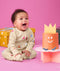 Infant baby wearing an ergoPouch Long Sleeve Romper 1.0 TOG Birthday Blobs, smiling widely at a cake