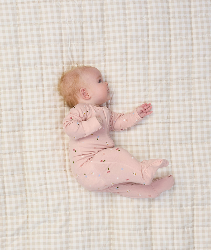 Baby lying on a beige checkered blanket, wearing a light pink ergoPouch Long Sleeve Romper with small floral prints.