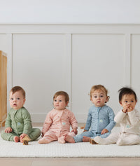 Four infants are seated side by side, each wearing ergPouch Long Sleeve Rompers in the colors of Willow, Daisies, Dragonflies, and Oatmeal Marle.