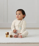 An infant is sitting dressed in an ergoPouch Long Sleeve Romper in Oatmeal Marle
