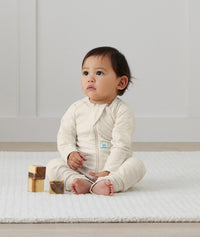 An infant is sitting dressed in an ergoPouch Long Sleeve Romper in Oatmeal Marle