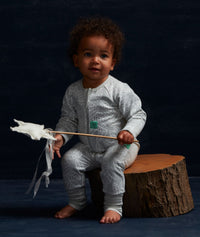 Baby wearing a light grey long sleeve romper with an all-over leaf print and front zip, sitting on a wooden log against a dark blue backdrop.