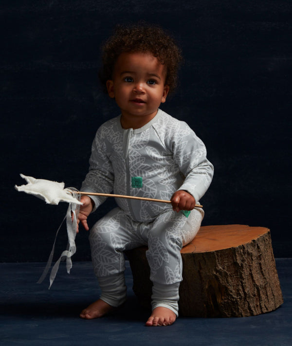 Baby wearing a light grey long sleeve romper with an all-over leaf print and front zip, sitting on a wooden log against a dark blue backdrop.