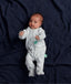 Baby lying on a dark navy blue fabric background, wearing a light grey, long-sleeved romper with a white leaf print.