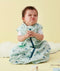Baby wearing a mint green sleep sack with a geometric pattern and front zipper, sitting on a soft surface with hands clasped and a playful scrunched facial expression.