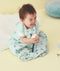 A smiling baby sits on a yellow floor wearing a light green, patterned sleep sack with dark blue chevron designs. The baby is laughing with their tongue out and holding a white block. The sleep sack features a front zipper and a snap at the bottom.