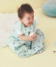 A smiling baby sits on a yellow floor wearing a light green, patterned sleep sack with dark blue chevron designs. The baby is laughing with their tongue out and holding a white block. The sleep sack features a front zipper and a snap at the bottom.