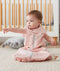 A baby sits on a light gray rug wearing a pink sleep sack with a white and gray leaf-like pattern. The baby is pointing off-camera, sitting in front of a white and wood crib. The sleep sack features a central gray zipper and a snap closure at the bottom.