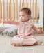 A baby sits on a light gray rug wearing a pink sleep sack with a white and gray leaf-like pattern. The baby is pointing off-camera, sitting in front of a white and wood crib. The sleep sack features a central gray zipper and a snap closure at the bottom.