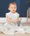 A baby sits on a light-colored blanket wearing a sleeveless, white sleep sack featuring a dark blue and yellow wavy pattern. The baby is holding a blue wooden toy camera and looking toward the camera against a dark blue background.