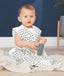 A baby sits on a light-colored blanket wearing a sleeveless, white sleep sack featuring a dark blue and yellow wavy pattern. The baby is holding a blue wooden toy camera and looking toward the camera against a dark blue background.