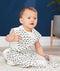 A baby sits on a light-colored blanket wearing a sleeveless, white sleep sack featuring a dark blue and yellow wavy pattern. 