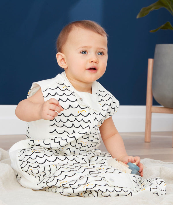 A baby sits on a light-colored blanket wearing a sleeveless, white sleep sack featuring a dark blue and yellow wavy pattern. 