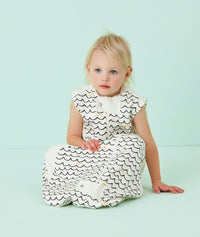 Infant wearing a white sleeveless sleep sack featuring a wave pattern, sitting against a light blue background.