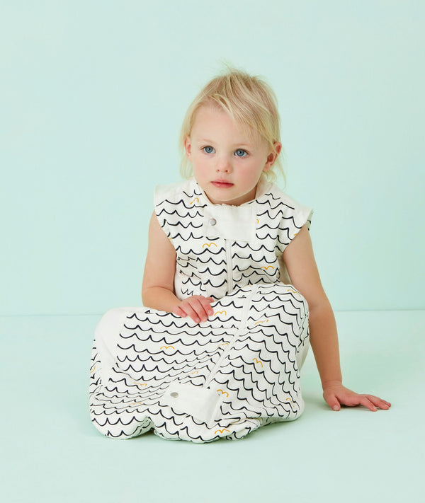 Infant wearing a white sleeveless sleep sack featuring a wave pattern, sitting against a light blue background.