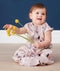 A baby sits on a light wood floor wearing a mauve sleep sack with an abstract pattern of black raindrops, white speckles, and peach-colored shapes. The baby is smiling and holding several yellow billy ball flowers against a dark blue background. The sleep sack has a full-length front zipper and a snap closure at the bottom.
