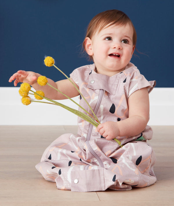 A baby sits on a light wood floor wearing a mauve sleep sack with an abstract pattern of black raindrops, white speckles, and peach-colored shapes. The baby is smiling and holding several yellow billy ball flowers against a dark blue background. The sleep sack has a full-length front zipper and a snap closure at the bottom.