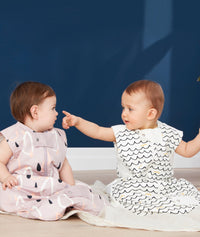 Two babies sit side-by-side on a light-colored blanket against a dark blue background. The baby on the left wears a mauve sleep sack with an abstract pattern of black raindrops and white and peach shapes. The baby on the right wears a white, sleeveless sleep sack with a black and yellow wavy pattern and points toward the other baby. Both sleep sacks feature a front zipper  and a snap closure at the bottom.