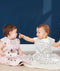 Two babies sit side-by-side on a light-colored blanket against a dark blue background. The baby on the left wears a mauve sleep sack with an abstract pattern of black raindrops and white and peach shapes. The baby on the right wears a white, sleeveless sleep sack with a black and yellow wavy pattern and points toward the other baby. Both sleep sacks feature a front zipper  and a snap closure at the bottom.