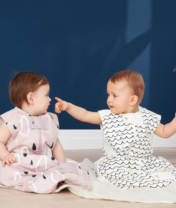 Two babies sit side-by-side on a light-colored blanket against a dark blue background. The baby on the left wears a mauve sleep sack with an abstract pattern of black raindrops and white and peach shapes. The baby on the right wears a white, sleeveless sleep sack with a black and yellow wavy pattern and points toward the other baby. Both sleep sacks feature a front zipper  and a snap closure at the bottom.
