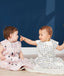Two babies sit side-by-side on a light-colored blanket against a dark blue background. The baby on the left wears a mauve sleep sack with an abstract pattern of black raindrops and white and peach shapes. The baby on the right wears a white, sleeveless sleep sack with a black and yellow wavy pattern and points toward the other baby. Both sleep sacks feature a front zipper  and a snap closure at the bottom.