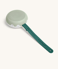 A sage green and white circular white noise machine with speaker holes on top and two buttons on the side sits attached to a dark green strap labeled “DRIFTAWAY ergoPouch®” on a white background.