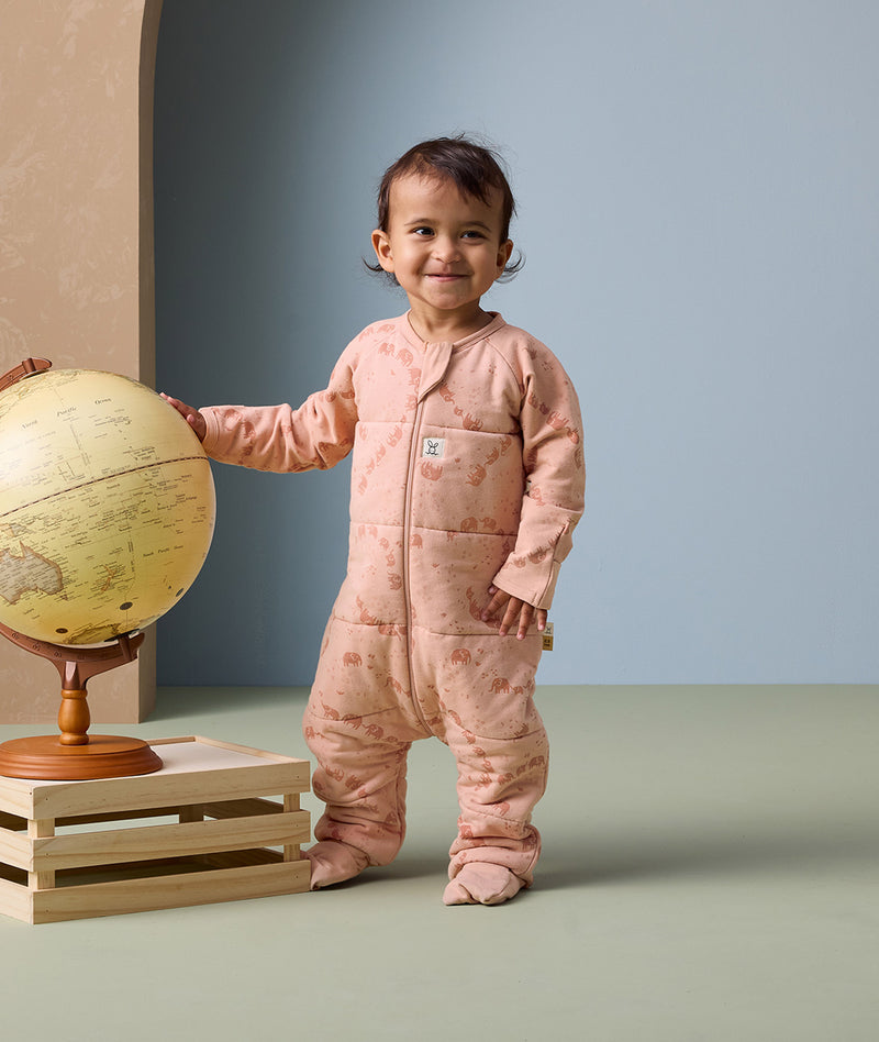 A toddler stands beside a globe, wearing an ergoPouch Sleep Onesie 2.5 TOG in Elephant Parade.