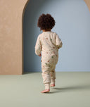 A toddler walks away from the camera wearing an ergoPouch Sleep Onesie with long sleeves and full-length legs. The onesie features an all-over animal print and a snug fit with foot cuffs, offering cozy comfort and flexibility for movement.