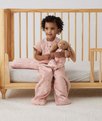 Toddler sitting on a crib in a Sleep Suit bag with her Organic Toddler Pillow next to her, all in Daisies print