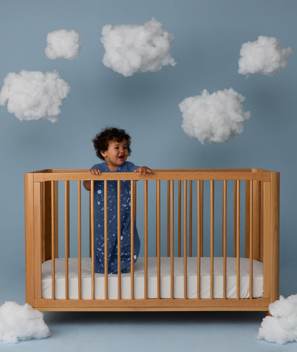 Child standing in a wooden crib wearing an ergoPouch Sleep Suit Sack 1.0 TOG Starbeam, a sleeping bag that converts to a sleep suit with legs.